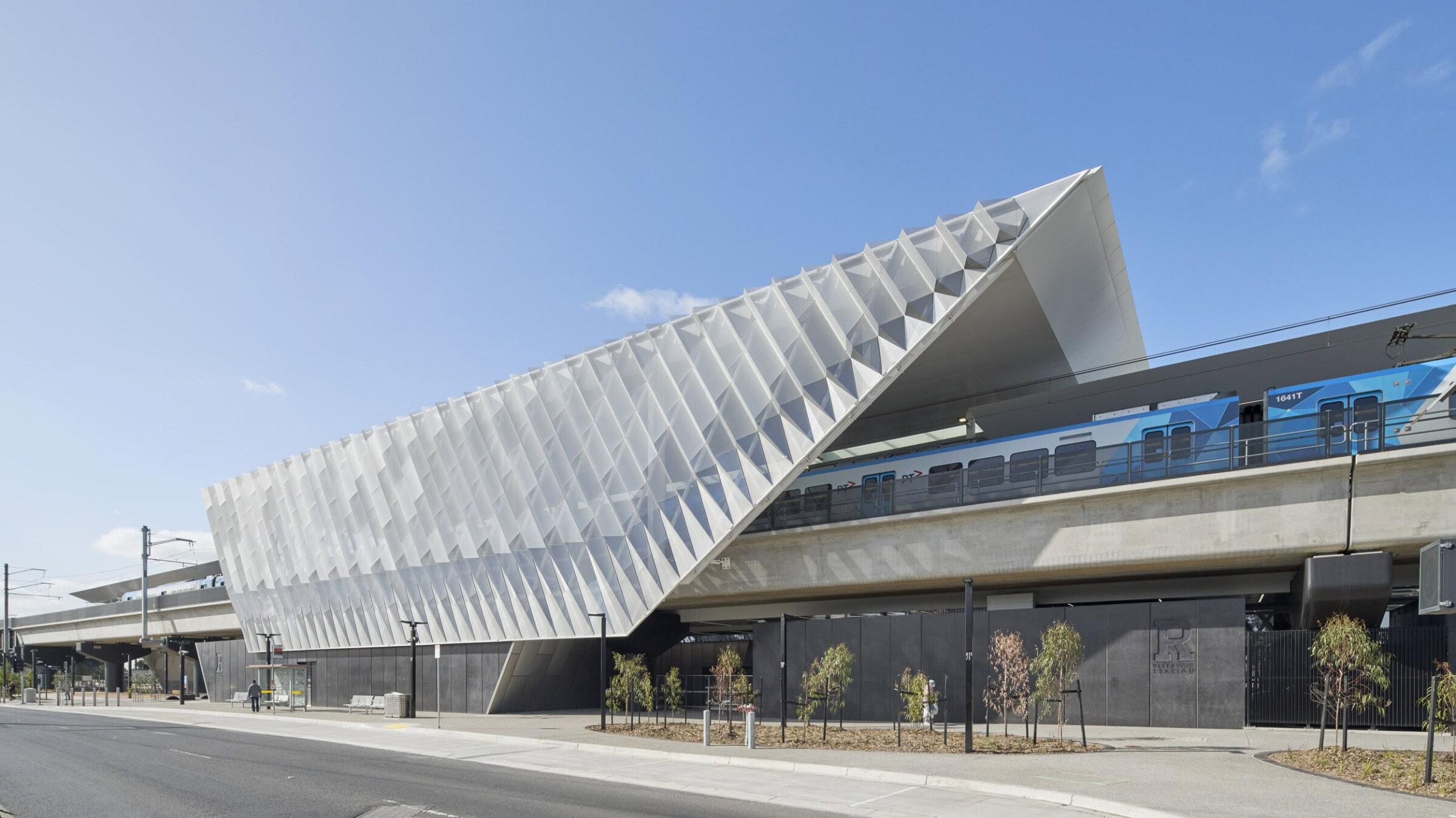 Elevated rail station at Reservoir with angular translucent canopy and passing Metro train – designed by Genton in Melbourne