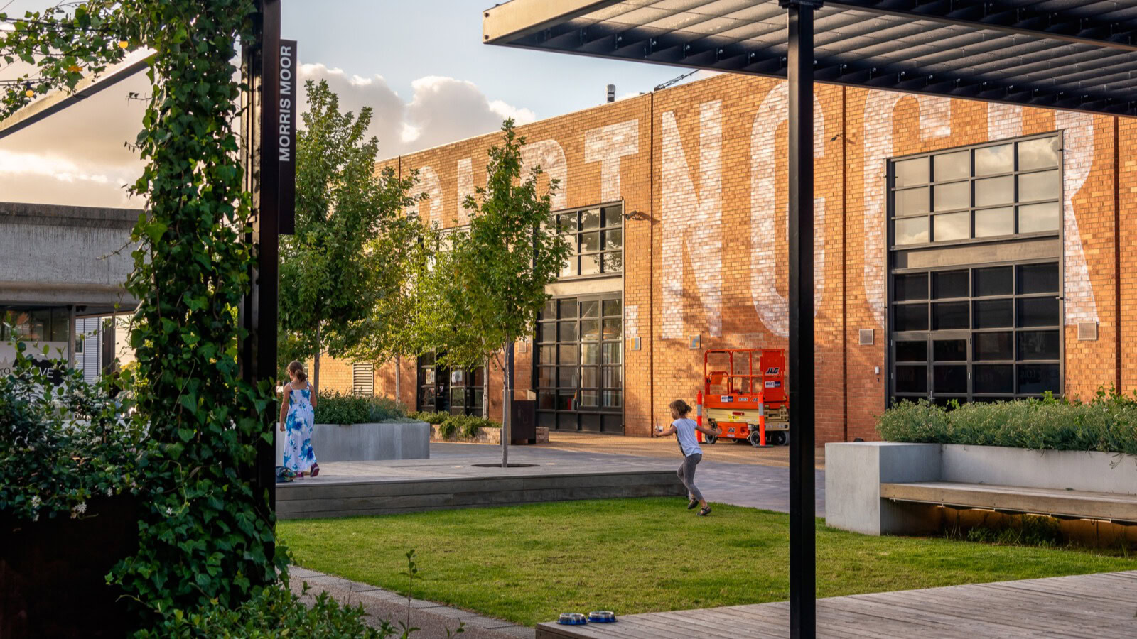 Children play on the grass in a landscaped courtyard at Morris Moor, a revitalised mixed-use precinct with adaptive reuse architecture and a bold brick façade.