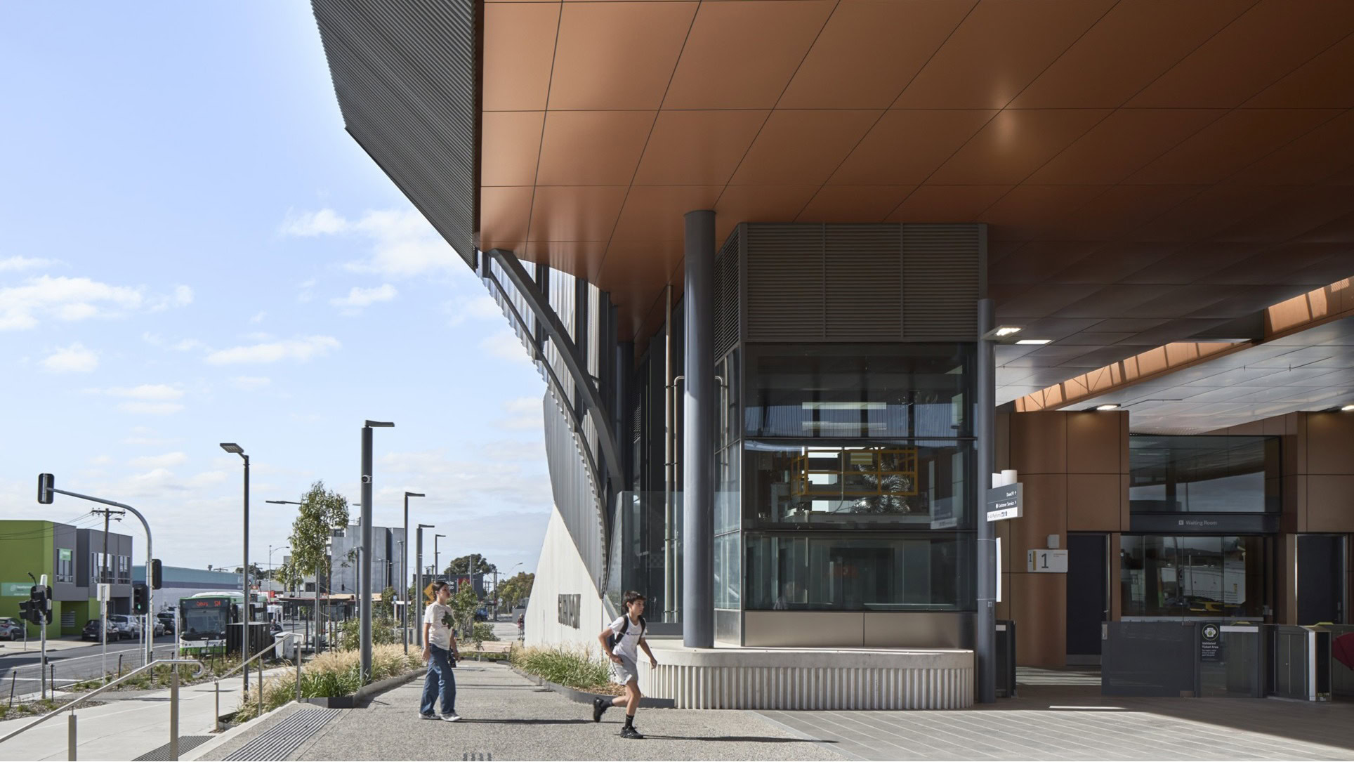 Glenroy Station Melbourne rail architecture with its copper-toned soffit and ticketing area at Glenroy Station, with pedestrians and surrounding urban context, designed by Genton,